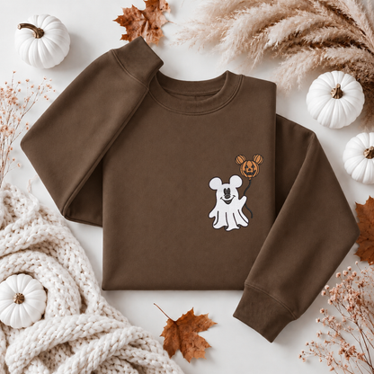 A flat-lay of a brown crewneck sweatshirt featuring a cute cartoon graphic of a white ghost and a jack-o'-lantern mouse. The sweatshirt is styled in an autumn-themed flat-lay with white pumpkins, dried leaves, pampas grass, and a chunky knit blanket on a white background.