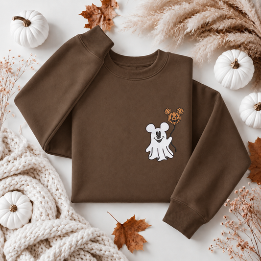 A flat-lay of a brown crewneck sweatshirt featuring a cute cartoon graphic of a white ghost and a jack-o'-lantern mouse. The sweatshirt is styled in an autumn-themed flat-lay with white pumpkins, dried leaves, pampas grass, and a chunky knit blanket on a white background.